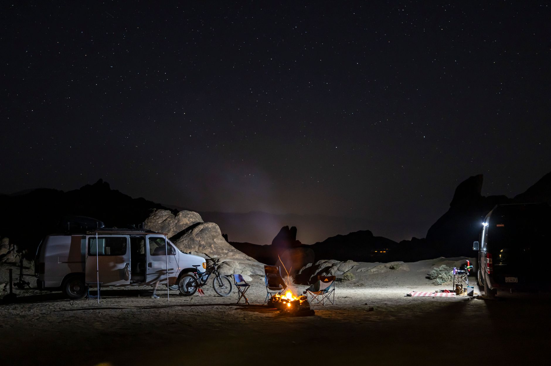 A desert camp after dark: a van, camp chairs, an e-bike and a fire under a full star field